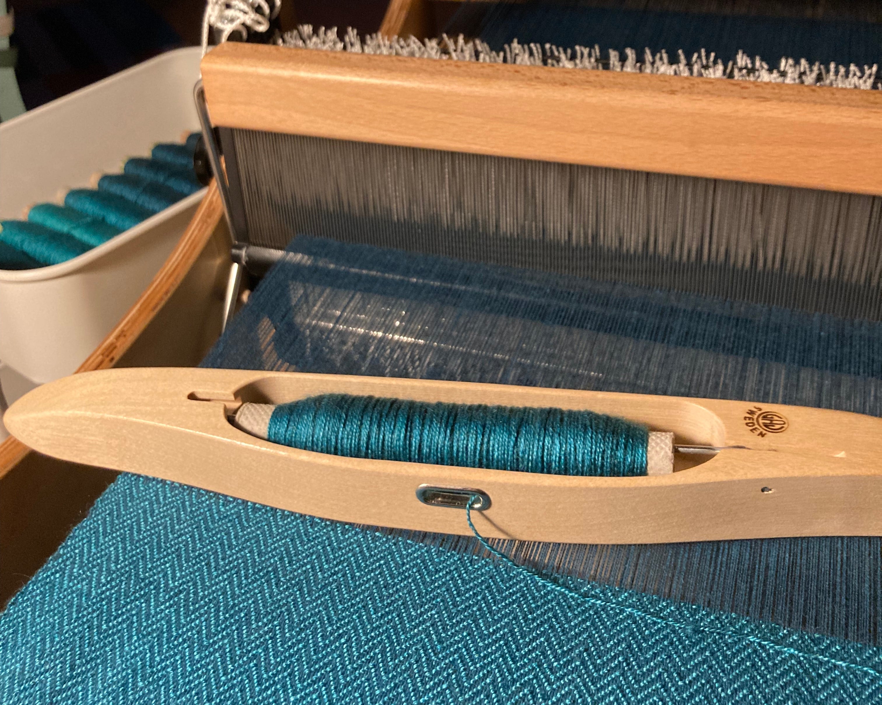 Louet Erica asztali szövőszéken készülő kék chevronmintás selymes  szövet vetélővel, háttérben felcsévélt fonalak 